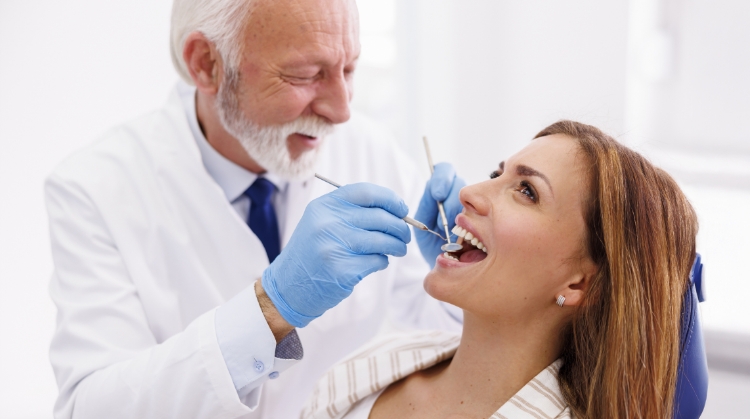 Smiling woman receiving a dental check-up from a senior male dentist wearing gloves, holding dental instruments in a clinic.